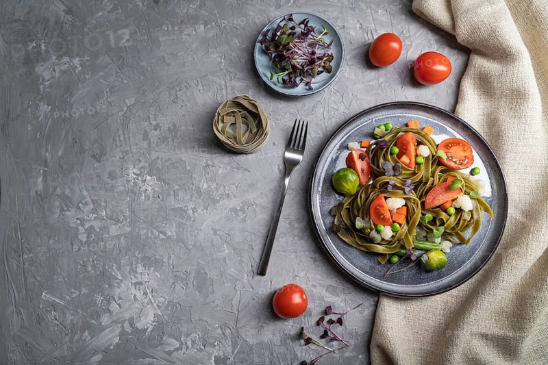 Tagliatelle green spinach pasta with tomato, pea and microgreen sprouts on a gray concrete background and linen textile. Top view, flat lay, copy space, hard light