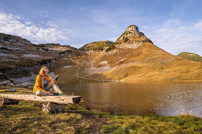 Lake Augstsee and the Atterkogel mountain on the Loser. A hiker sits on a bench and reads a book. Autumn, good weather, blue sky. Altaussee, Bad Aussee, Ausseer Land, Totes Gebirge, Styria, Upper Austria, Austria, Europe