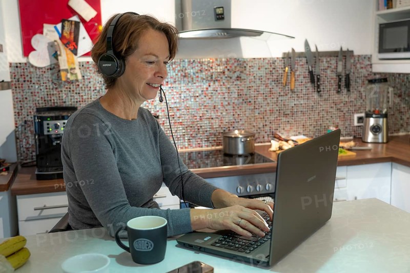 Woman, mid-50s, works from home, with laptop and communicates with colleagues via headset, home office, in the kitchen