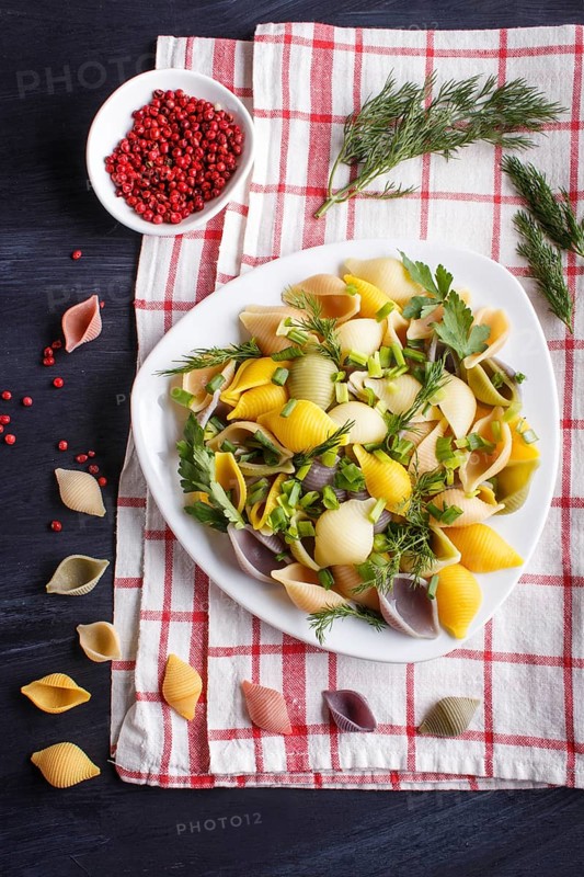 Conchiglie colored pasta with fresh greengrocery on a linen tablecloth on black wooden background. top view, flat lay