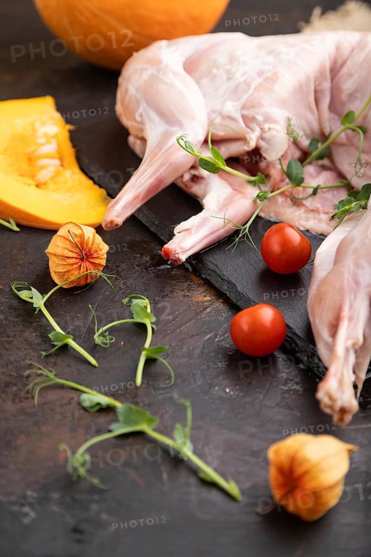 Whole raw rabbit with pumpkin, tomatoes, pea sprouts on a black concrete background and linen textile. Side view, close up, selective focus