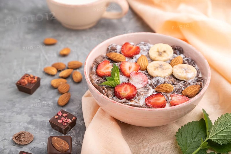 Chocolate cornflakes with milk, strawberry and almonds in ceramic bowl on gray concrete background and orange linen textile. Side view, close up, selective focus