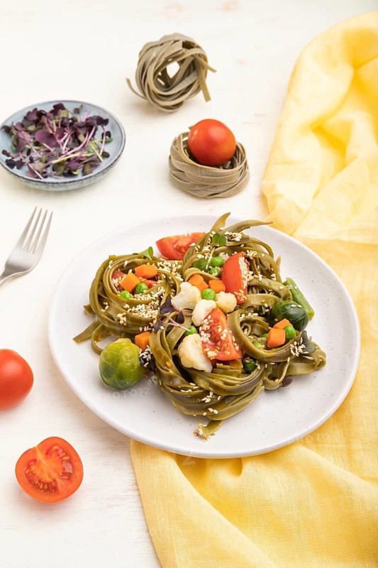 Tagliatelle green spinach pasta with tomato, pea and microgreen sprouts on a white wooden background and yellow textile. Side view, close up