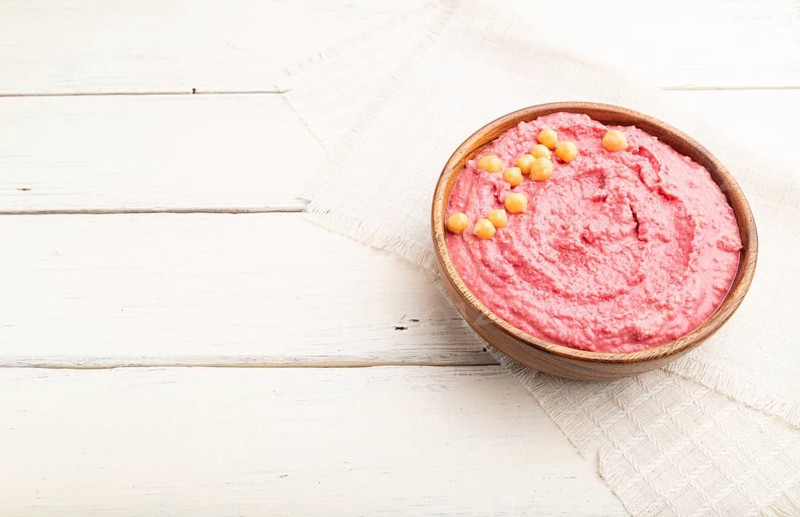 Hummus with beet in wooden bowl on a white wooden background and linen textile. Side view, copy space