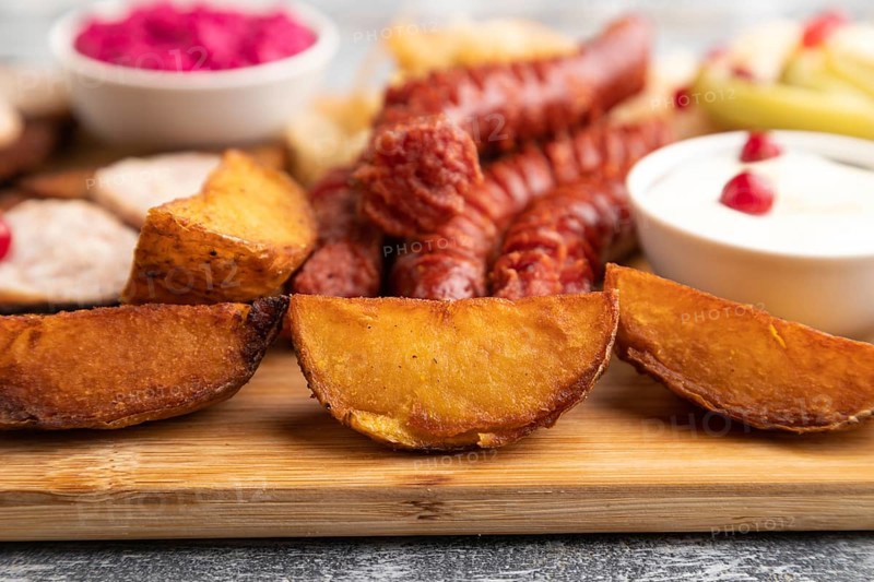 Set of snacks: sausages, toast, sauerkraut, marinated onion and cucumber, baked potato on a cutting board on a gray wooden background. Side view, close up, selective focus