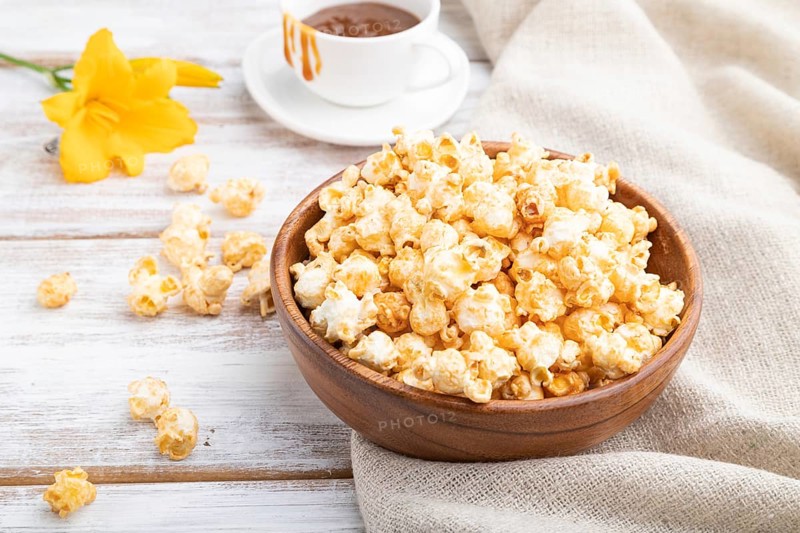 Popcorn with caramel in wooden bowl and a cup of coffee on a white wooden background and linen textile. Side view, close up
