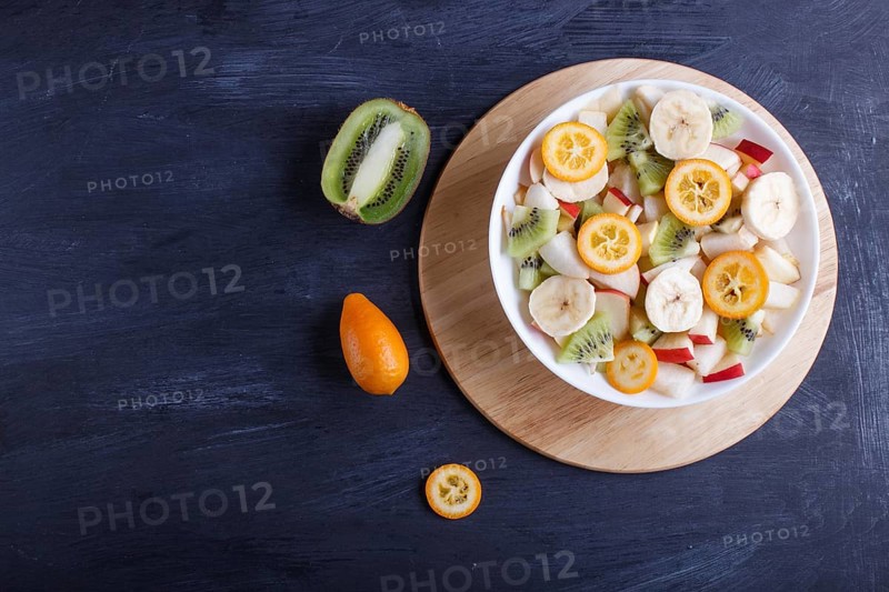 Vegetarian salad of bananas, apples, pears, kumquats and kiwi on black woodem background, top view, copy space