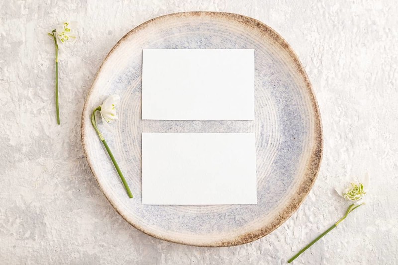 White paper invitation card, mockup with galanthus snowdrop flowers on ceramic plate and gray concrete background. Blank, flat lay, top view, still life, copy space, wedding invitation