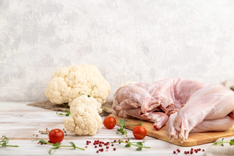 Whole raw rabbit with cauliflower, tomatoes and spices on a white wooden background and linen textile. Side view, copy space
