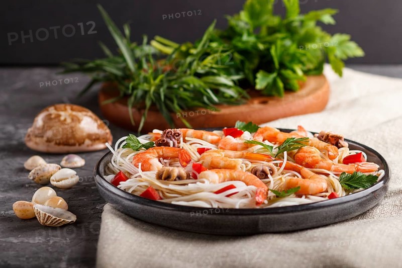Rice noodles with shrimps or prawns and small octopuses on gray ceramic plate on a black concrete background and linen textile. side view, close up, selective focus