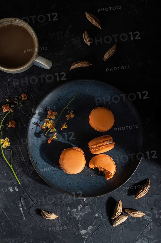Orange macarons or macaroons cakes with cup of coffee on a black concrete background. Hard light, contrast, low key. Top view, flat lay, close up