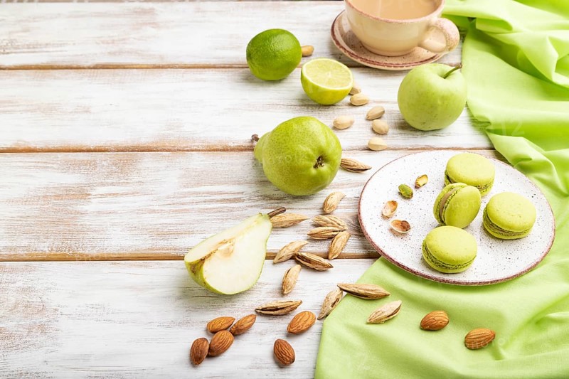 Green macarons or macaroons cakes with cup of coffee on a white wooden background and green linen textile. Side view, copy space