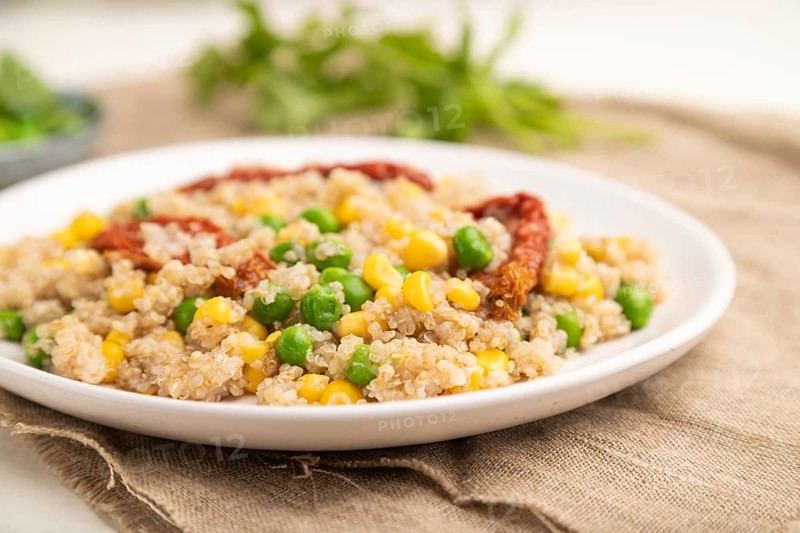 Quinoa porridge with green pea, corn and dried tomatoes on ceramic plate on a white wooden background and linen textile. Side view, close up, selective focus