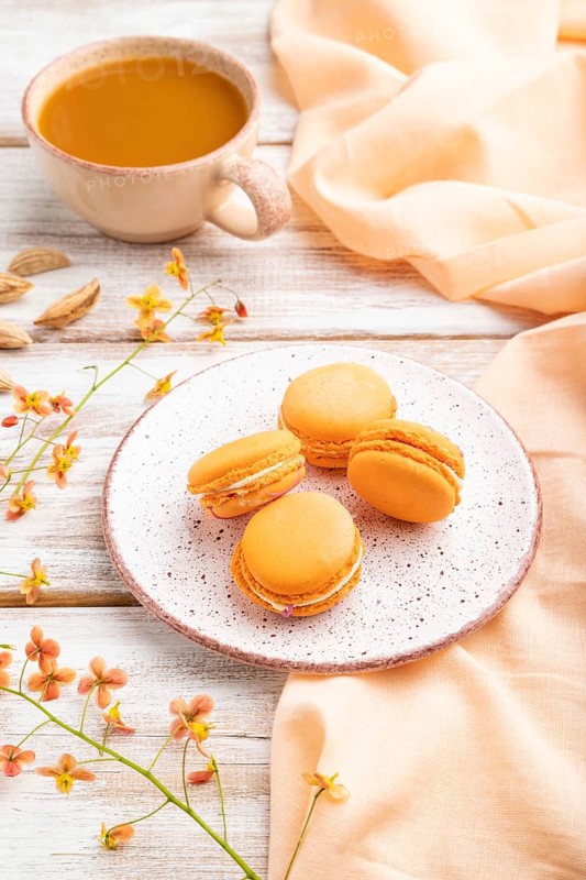 Orange macarons or macaroons cakes with cup of apricot juice on a white wooden background and orange linen textile. Side view, close up