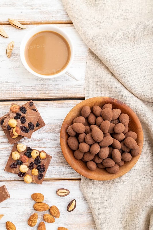 Almond in chocolate dragees in wooden plate and a cup of coffee on white wooden background and linen textile. Top view, close up, flat lay