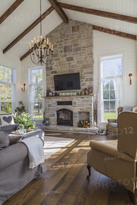 Dark grey upholstered ektorp model sofas, bergere armchairs, tan, beige and grey nuanced natural cut stone wood burning fireplace in living room with rug and stained oak wood floorboards inside country style home, Quebec, Canada, North America