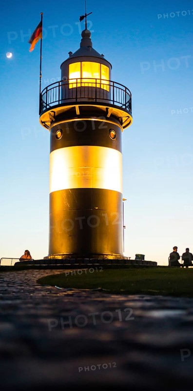 Black and white lighthouse, called 'Kleiner Preuße', stands illuminated in the evening against a clear blue sky, bright beacon, rising moon, German flag, silhouettes, people sitting on benches, paved curved path in the foreground, frog perspective, mouse perspective, twilight, evening, dusk, calm atmosphere, North Sea resort Wremen, Lower Saxony Wadden Sea National Park, World Heritage Site, Wurster North Sea coast, Land Wursten, Cuxhaven district, Lower Saxony, Germany, Europe