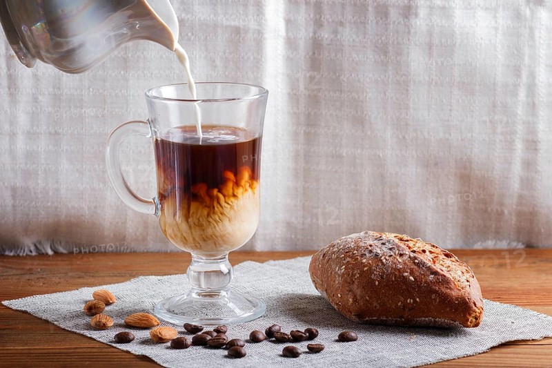 Glass cup of coffee with cream poured over and bun on a wooden background and linen textile. close up, copy space