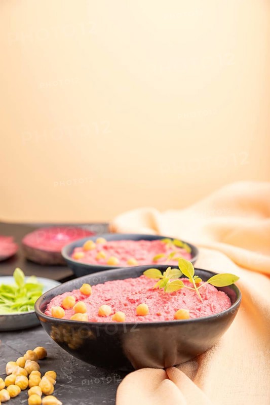 Hummus with beet and microgreen basil sprouts in ceramic bowl on a black concrete background and orange textile. Side view, copy space, selective focus