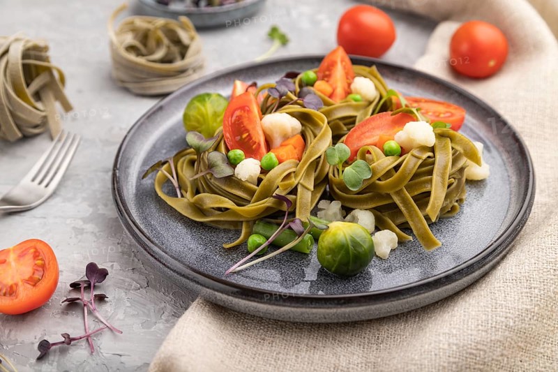 Tagliatelle green spinach pasta with tomato, pea and microgreen sprouts on a gray concrete background and linen textile. Side view, close up, selective focus