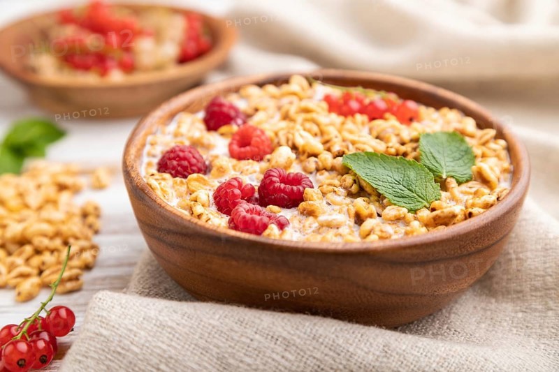Wheat flakes porridge with milk, raspberry and currant in wooden bowl on white wooden background and linen textile. Side view, close up, selective focus