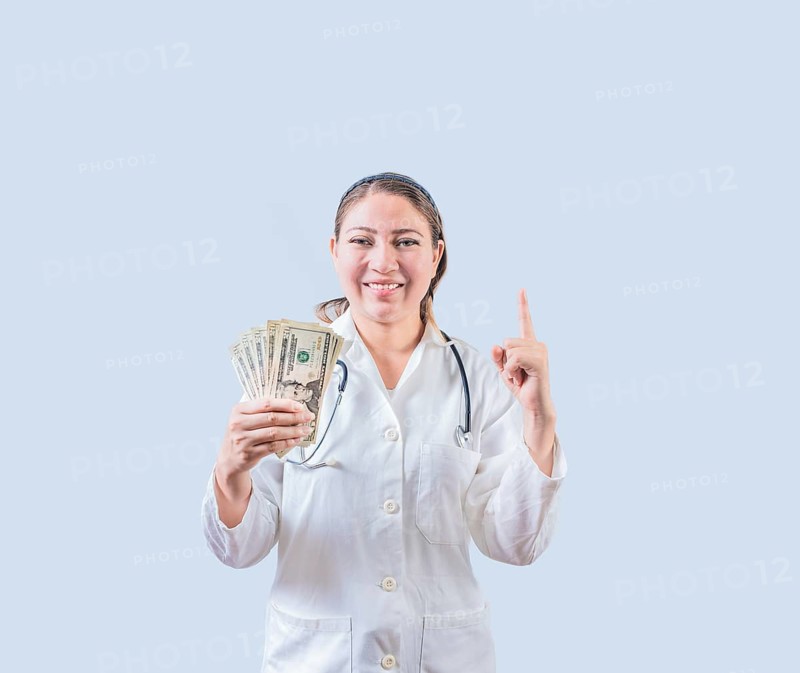 Smiling female doctor holding money and pointing up isolated. Happy young female doctor showing money and pointing at promo up