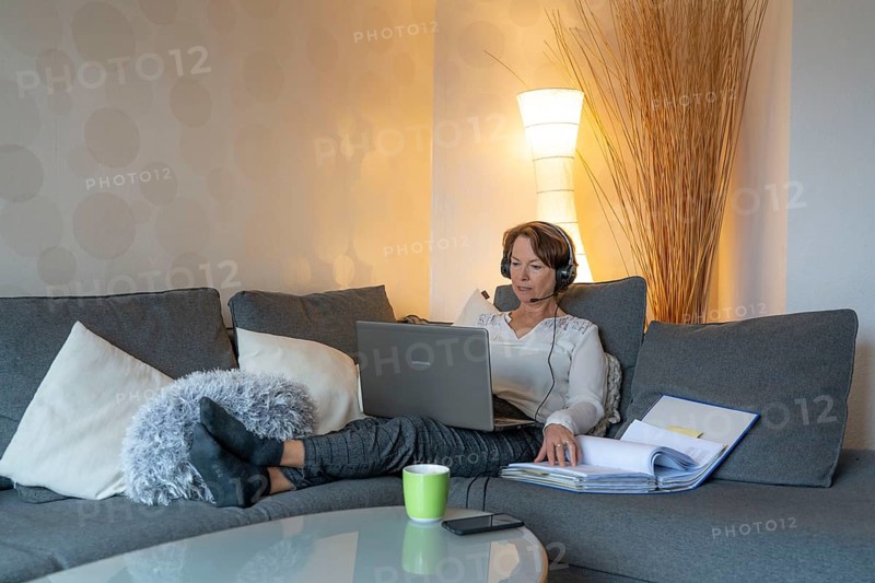 Woman, mid-50s, works from home, with laptop and communicates with colleagues via headset, home office, on the sofa
