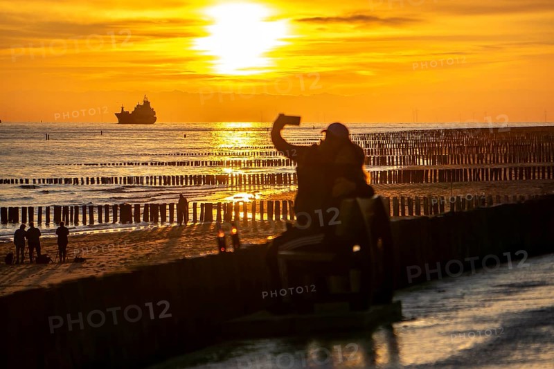 Sunset on the beach of Zoutelande, beach with wooden pile breakwaters, tourists, cargo ship sailing towards the Westerschelde, Zeeland, Netherlands