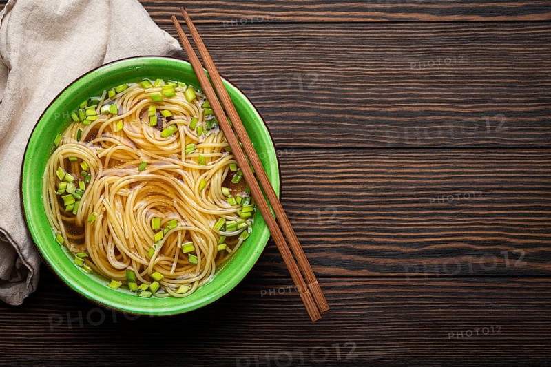 Asian noodles soup in green rustic ceramic bowl with wooden chopsticks top view on dark wooden background. Lo mein noodles with bouillon and green onion, space for text, food photography