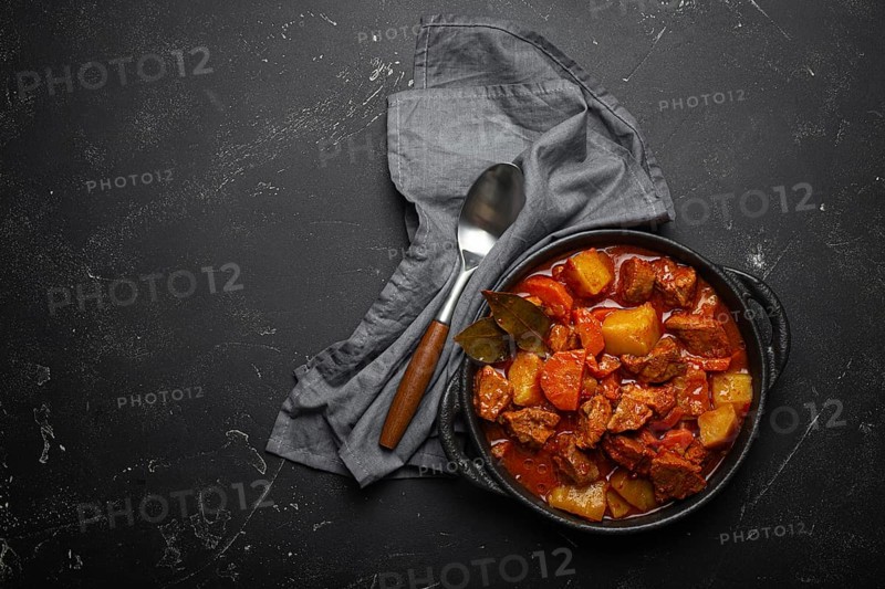 Beef meat stew with potatoes, carrot and delicious gravy in black casserole pot with bay leaves with spoon on black dark rustic concrete background from above, space for text, food photography
