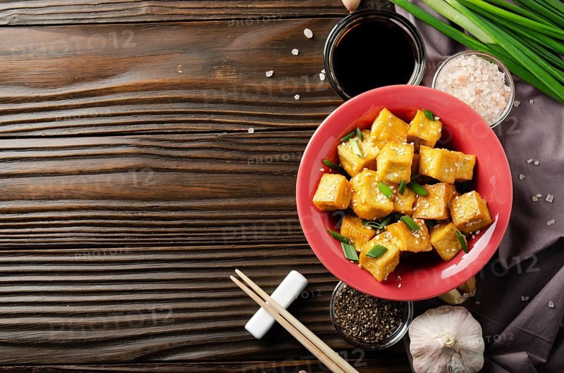 Flat lay view at stir fried tofu cubes with chives in clay dish on wooden kitchen table with napkin chives pepper garlic and soy sauce aside