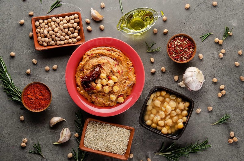 Hummus topped with chickpeas, olive oil and sun dried tomato on stone table with different spices aside. Flat lay
