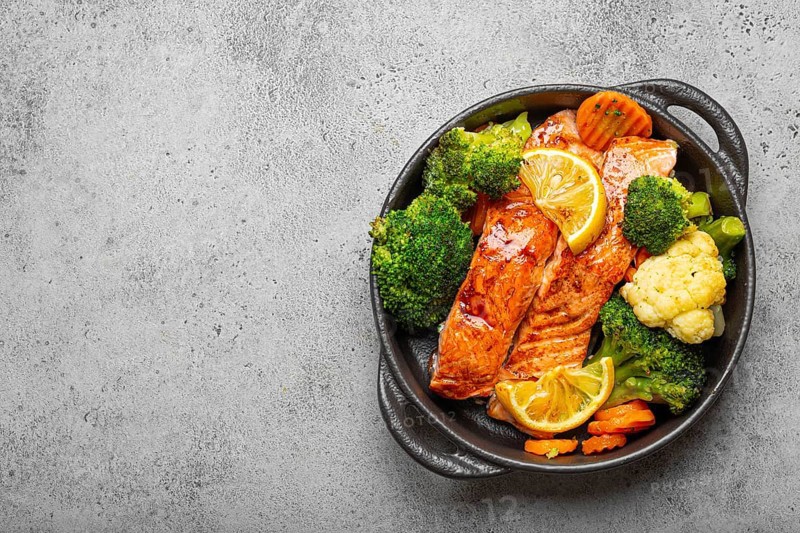 Healthy baked fish salmon steaks, broccoli, cauliflower, carrot in black cast iron casserole bowl on grey stone background. Cooking a delicious low carb dinner, healthy nutrition, space for text, food photography