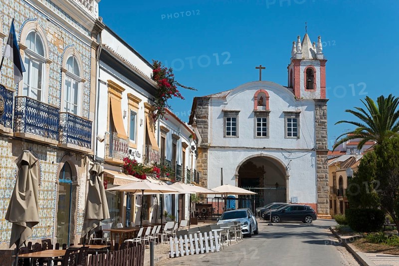 Street view with cafés, a white and red church and lush vegetation under a bright blue sky, house with azulejos on the left, Igreja de Nossa Senhora da Ajuda ou de São Paulo, Church of Our Lady of Help or São Paulo, Praça Dr António Padinha, Tavira, Faro, Algarve, Portugal, Europe