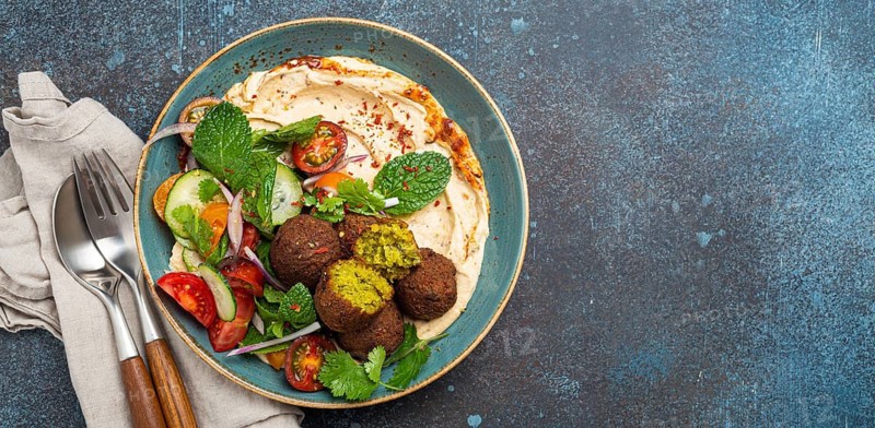 Middle Eastern Arab meal with fried falafel, hummus, vegetables salad with fresh green cilantro and mint leaves in ceramic bowl on stone rustic background top view. Arabic cuisine, space for text, food photography