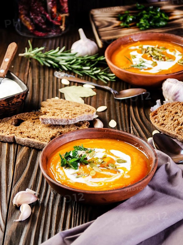 Homemade rustic pumpkin soup with seeds in clay dish on wooden table with bread spices and greens aside