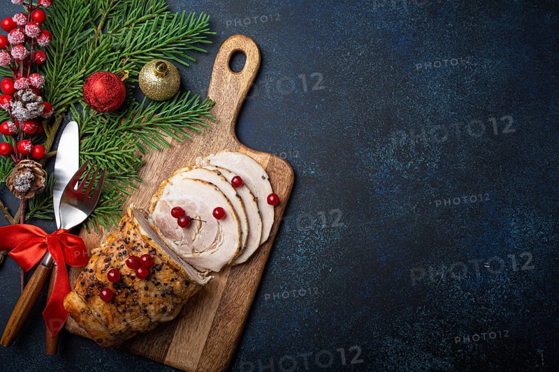 Christmas baked ham sliced with red berries and festive decorations on wooden cutting board, dark rustic background from above. Christmas and New Year holiday dinner with baked pork, space for text, food photography