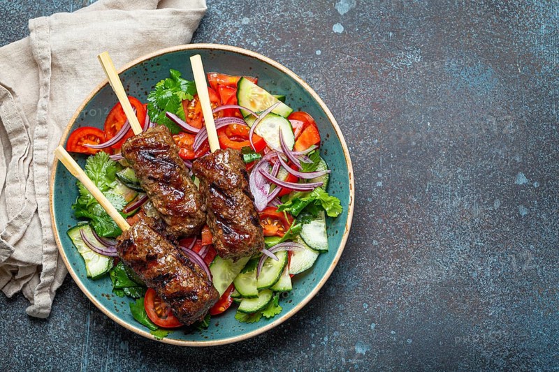 Grilled skewer meat beef kebabs on sticks served with fresh vegetables salad on plate on rustic concrete background from above. Traditional Middle Eastern and Turkish dish Kebab, space for text, food photography