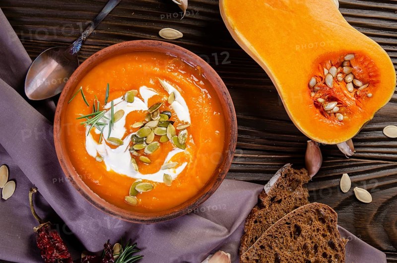 Flat lay view at homemade rustic pumpkin soup with seeds in clay dish on wooden table with bread spices and greens aside