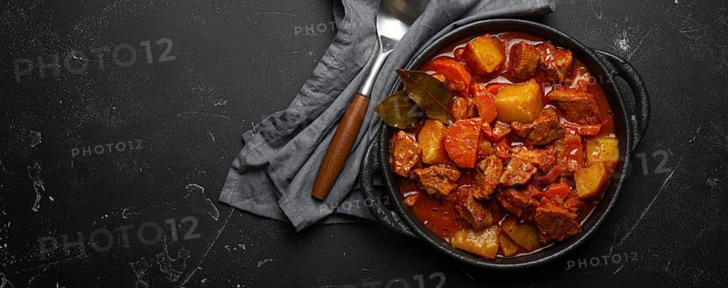 Beef meat stew with potatoes, carrot and delicious gravy in black casserole pot with bay leaves with spoon on black dark rustic concrete background from above, space for text, food photography