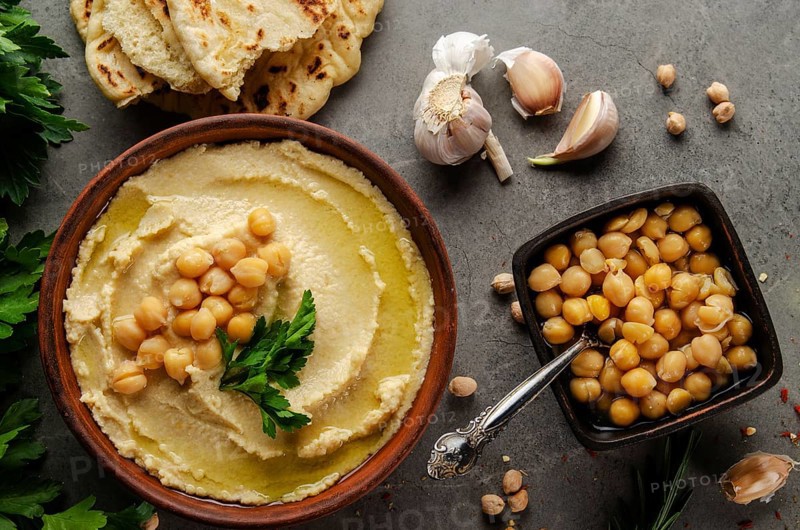 Hummus topped with chickpeas, olive oil and green coriander leaves on stone table with pita bread aside
