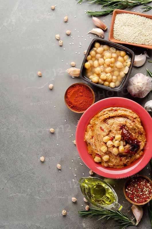 Hummus topped with chickpeas, olive oil, green coriander leaves and sun dried tomatoes on stone table with pita bread and spices aside. Flat lay