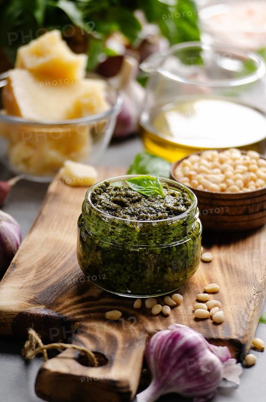 Glass jar with genovese pesto sauce on cutting board with basil leaves, pine nuts, parmesan cheese, olive oil and garlic aside