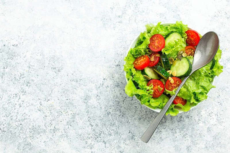 Healthy vegetables salad with fresh cucumbers, tomatoes, fresh iceberg salad leaves in a bowl on rustic white concrete table top view with a spoon. Veggie salad for diet concept, space for text, food photography