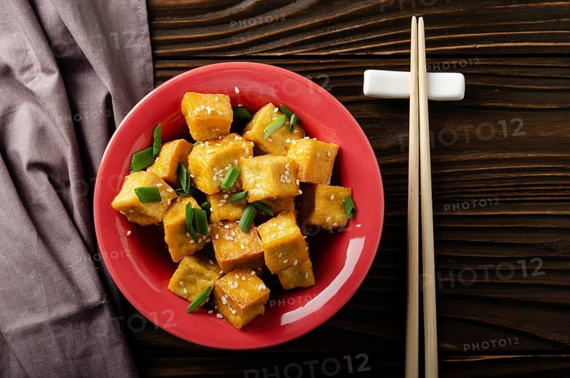 Flat lay view at crispy deep stir fried tofu cubes with chives in clay dish on wooden kitchen table with napkin aside