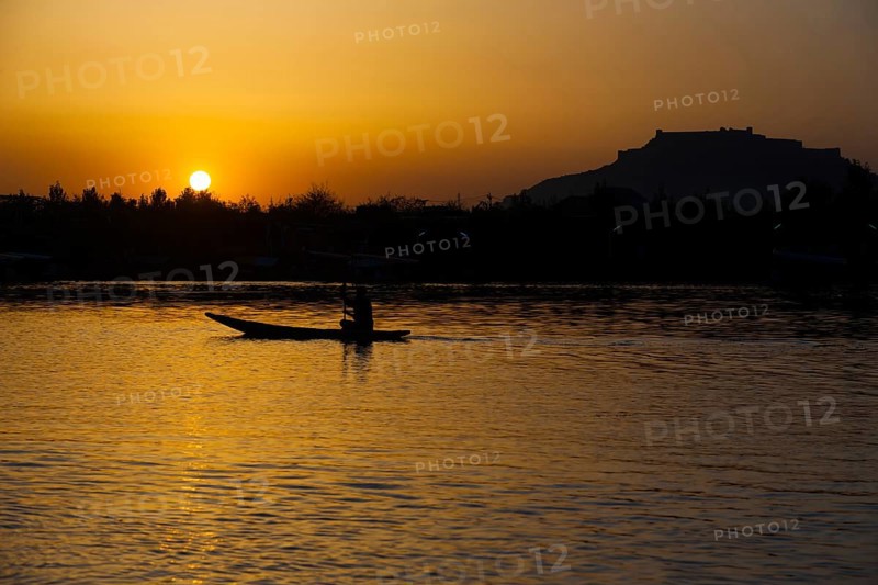The silhouette of a boat glides along the surface of Dal Lake during sunset with Srinagar Fort in the background in Kashmir, India, Asia