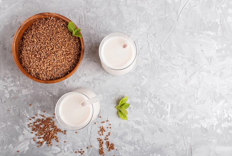 Organic non dairy buckwheat milk in glass and wooden plate with buckwheat seeds on a gray concrete background. Vegan healthy food concept, flat lay, top view, copy space
