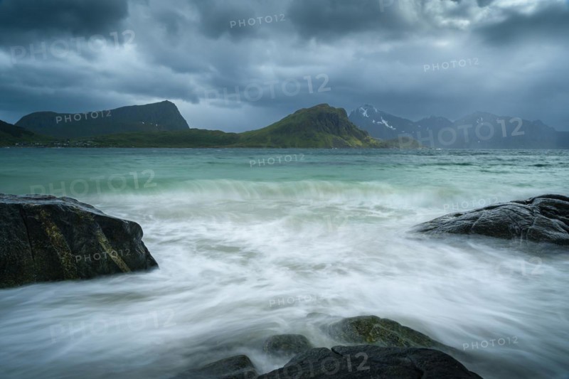 Seascape on the beach at Haukland. View of the mountains of Vestvagoya and Myrland on Flakstadoya. Offersoykammen on the left. Clouds in the sky, some sun. Rocks in the foreground. A wave in the sea. Early summer. Long exposure. Haukland Beach, Haukland, Vestvagoya, Lofoten, Norway, Europe