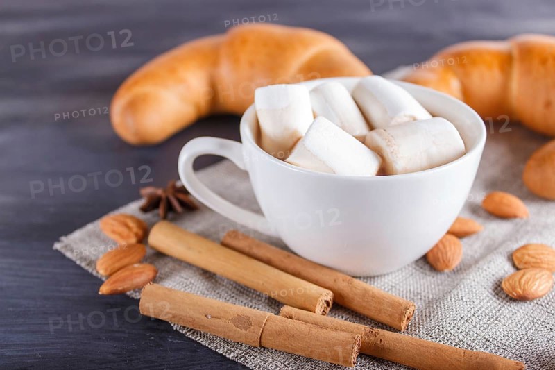 A cup of hot chocolate with marshmallow, croissants and spices on linen napkin and black wooden background. close up, copy space