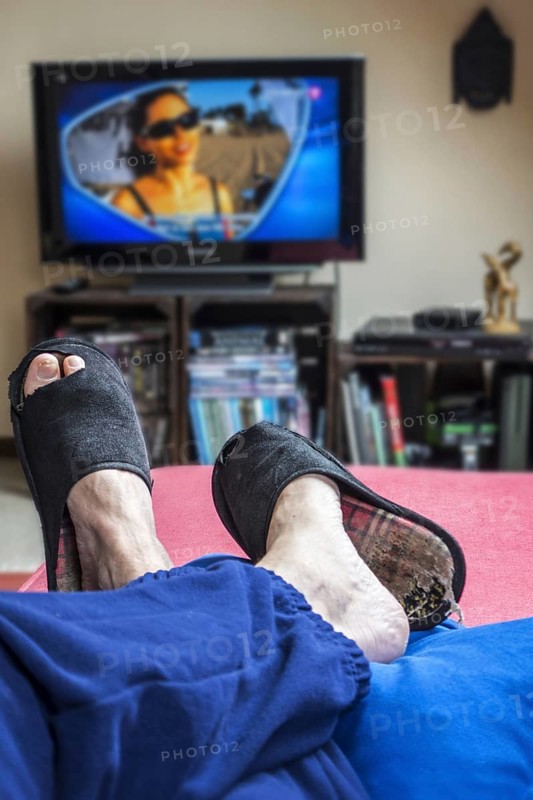 Couch potato, lazy man in comfy chair wearing worn slippers with big toes sticking through and watching television set in living room